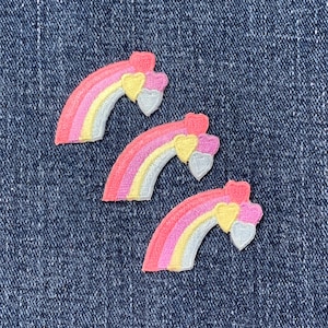 May include: Three embroidered rainbow patches with pink, yellow, and silver arches, topped with heart-shaped accents. The patches are set against a blue denim background, creating a colorful and cheerful design.
