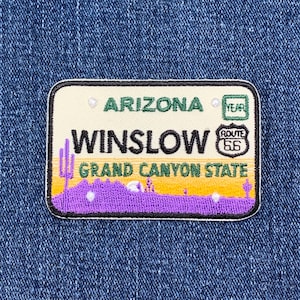 May include: Embroidered patch resembling an Arizona license plate. The patch features the text "ARIZONA", "WINSLOW", and "GRAND CANYON STATE". The design includes a Route 66 logo and a purple and yellow landscape.