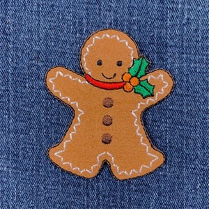 May include: A brown gingerbread man patch with white stitching, a red scarf, and a sprig of holly. The patch has three brown buttons and a smiling face. The patch is on a blue denim background.