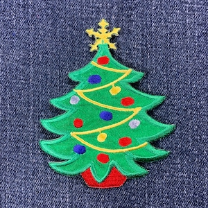 Könnte beinhalten: Gestickter Weihnachtsbaum-Aufnäher mit einem grünen Baum, roter Basis und gelbem Stern. Der Baum ist mit bunten Ornamenten und gelben Lichterketten geschmückt. Der Aufnäher befindet sich auf einem blauen Jeans-Hintergrund.