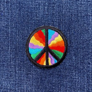May include: Embroidered peace sign patch with a black border and a rainbow-colored design. The patch is circular and sewn onto a blue denim fabric.