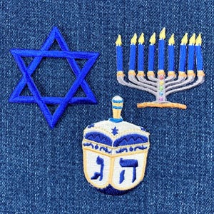 May include: Blue embroidered Star of David, a blue and white dreidel, and a menorah with nine blue candles on a denim background.