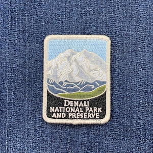May include: A rectangular embroidered patch featuring a light blue sky above a white and grey mountain range, a dark blue valley, and a green foreground. The bottom black section contains the white text DENALI NATIONAL PARK AND PRESERVE.