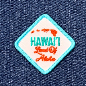 May include: A diamond-shaped patch with a white background and a turquoise border. The patch features the word "HAWAII" in turquoise, the phrase "Land Of Aloha" in orange, and an orange outline of the Hawaiian islands.