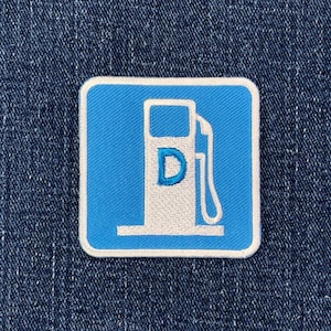 May include: A square patch featuring a blue background with a white border. The patch displays a white gas pump icon with the letter "D" on it. The patch is set against a blue denim background.