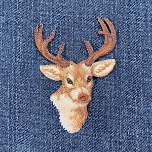 May include: Embroidered patch featuring a deer head with detailed antlers, ears, and facial features. The patch is primarily tan and brown, with a white outline, set against a blue denim background. Ideal for adding to clothing or accessories.