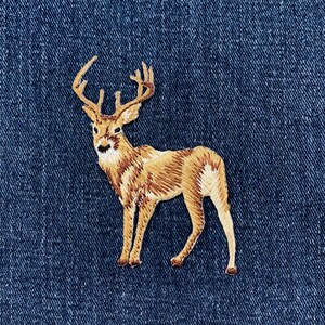 May include: An embroidered patch featuring a standing deer with large antlers, rendered in shades of tan, brown, and cream thread. The deer is depicted in profile, facing left, with detailed stitching that creates a textured appearance on a dark blue denim fabric.