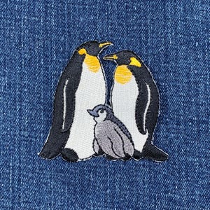 May include: Embroidered patch depicting a penguin family. Two adult penguins with black and white bodies and yellow details stand beside a grey baby penguin. The patch is on a blue denim background.