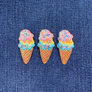 May include: Three embroidered ice cream cone patches. Each patch has a glittery, multi-layered ice cream scoop in pink, yellow, and blue, with iridescent sequins. The cones are a textured, golden-brown colour. The patches are set against a blue denim background.