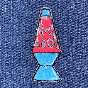 Könnte beinhalten: Ein Aufnäher mit einer Lavalampe mit glitzerndem silbernen Innenraum und roten, rosa und blauen Details. Der Aufnäher ist schwarz umrandet und befindet sich auf einem blauen Jeanshintergrund. Das Lavalampe-Design ist ein Accessoire im Retro-Stil.