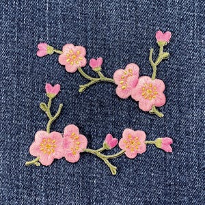 May include: Two embroidered floral patches featuring pink cherry blossoms with yellow centres and green stems. The patches are arranged on a blue denim background, ideal for adding a decorative touch to clothing or accessories.