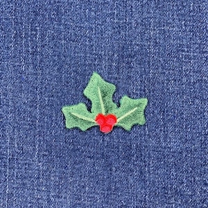 May include: A green embroidered holly leaf with red berries, set against a blue denim background. The holly design is detailed with light green stitching, creating a festive and seasonal embellishment.