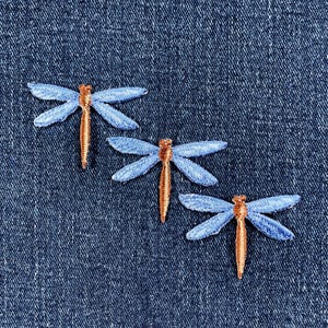 May include: Three embroidered dragonfly patches. Each dragonfly has light blue wings and a copper-colored body. The patches are on a dark blue denim background.