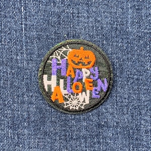 May include: A round, green, glittery patch with a white spiderweb design. The words "Happy Halloween" are embroidered in purple and white letters around a cartoon orange pumpkin with a black face.