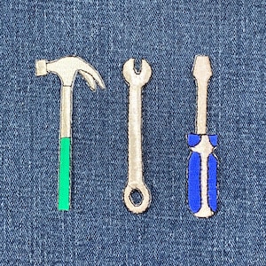 May include: Three embroidered tool patches: a hammer with a green handle, a wrench, and a screwdriver with a blue handle. The patches are silver and white, and are set against a blue denim background.