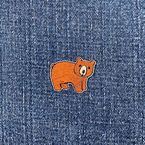 May include: A small, embroidered patch featuring a cartoon bear in shades of orange and brown, outlined in white. The patch is set against a textured, dark blue denim background.