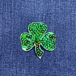 May include: A green sequined shamrock applique on a blue denim background. The shamrock is made of small, shiny green sequins and has a white border. The three-leaf clover is a symbol of good luck.