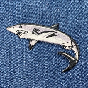 May include: An embroidered shark patch in white and grey with black detailing. The shark is swimming to the right with its mouth open.