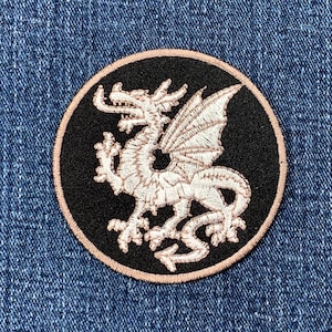 May include: Circular embroidered patch with a white dragon design on a black background. The patch has a light pink border and is displayed on a blue denim surface. The dragon has detailed wings and scales.