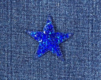 May include: A close-up of blue denim fabric featuring a sparkling, blue sequined star. The star is centrally placed and has a five-point design. The denim has a textured appearance.