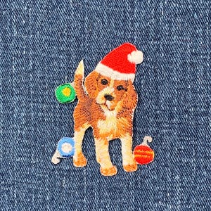 以下が含まれることがあります： 赤いサンタ帽をかぶった茶色と白の犬の刺繍パッチ。犬は緑、青、赤などカラフルなクリスマスオーナメントに囲まれています。青いデニムの背景にあります。
