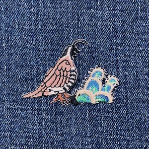 以下が含まれることがあります： 黒い頭、ピンクと白の体、青と緑の花柄が特徴の鳥の刺繍パッチ。 パッチは青いデニムの背景に配置されています。 衣類やアクセサリーに装飾的なタッチを加えるのに最適です。 長さは約5cmです。