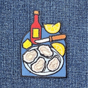 May include: Embroidered patch featuring a plate of oysters, lemon wedges, a red bottle, and an oyster knife. The patch has a blue background and a black border. The oysters are beige, and the lemons are yellow.