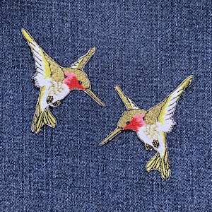 以下が含まれることがあります： 2つの刺繍されたハチドリのパッチ。各パッチは、赤い喉、白い胸、金と黄色の羽を持つハチドリが特徴です。パッチは青いデニムの背景に配置されています。
