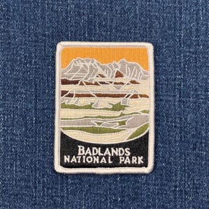 May include: Embroidered patch depicting a landscape with mountains and a sunset. The rectangular patch has a white border and the text "BADLANDS NATIONAL PARK". The patch is set against a blue denim background.