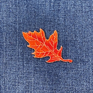 May include: An orange embroidered leaf patch with white stitching details, set against a blue denim background. The leaf design is a fall theme, perfect for adding to clothing or accessories.