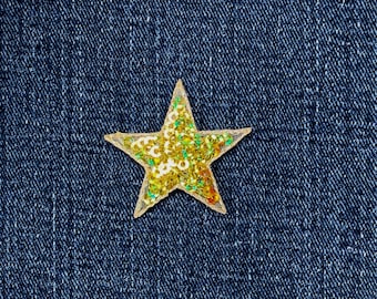 May include: A gold star-shaped sequin patch with a clear border, filled with gold and green sequins. The patch is set against a dark blue denim background.