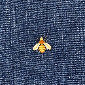May include: A small, embroidered bee patch with orange, yellow, and white details. The bee is centered on a textured, dark blue denim background. The patch is likely for clothing or accessories.