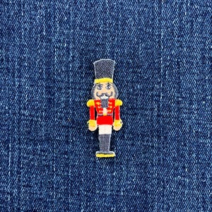 Könnte beinhalten: Ein gestickter Aufnäher in Form eines Nussknackers mit hohem schwarzem Hut, roter Jacke mit gelben Akzenten und grauer Hose. Die Figur hat ein weißes Gesicht mit schwarzem Schnurrbart und ist auf einem dunkelblauen Jeansstoff platziert.