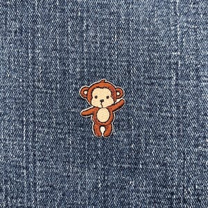 May include: A small, embroidered patch featuring a cartoon monkey in shades of brown and tan. The monkey is waving with one arm raised. The patch is set against a textured, dark blue denim background.
