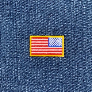 May include: A small embroidered patch depicting the American flag. The patch has a yellow border, red and white stripes, and a blue field with white stars. It is set against a blue denim background.