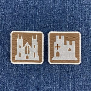 Könnte beinhalten: Zwei quadratische, bestickte Aufnäher mit abgerundeten Ecken. Der linke Aufnäher zeigt die Silhouette einer hellgrauen Kathedrale auf braunem Grund. Der rechte Aufnäher zeigt die Silhouette einer hellgrauen Burg mit einem kleinen braunen Kreuz.