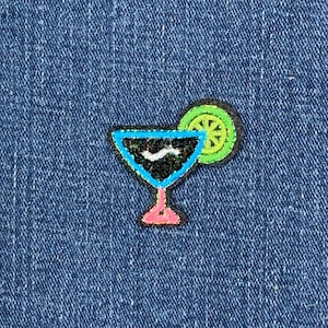 May include: A cocktail patch featuring a black drink, a lime slice, and a pink stem. The cocktail glass has a blue outline and a black interior. The lime slice is green with yellow details. The patch is on a blue denim background.