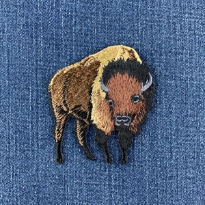 May include: An embroidered patch featuring a bison in shades of brown, tan, and black. The bison is depicted in a standing position, showcasing detailed stitching that highlights its thick fur, curved horns, and facial features against a blue denim fabric.