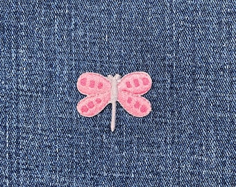 May include: A pink embroidered butterfly patch with detailed wing patterns. The patch is set against a textured blue denim background, creating a contrast in colour and texture. The butterfly's wings have a darker pink outline.