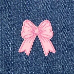 May include: A pink bow-shaped patch with a detailed design. The patch is set against a blue denim background. The bow has a detailed outline and is a solid, soft pink colour.
