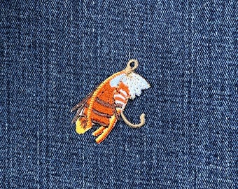 May include: Embroidered patch of a stylized insect with orange, white, and yellow details. The patch has a loop at the top and a curved hook shape. The patch is on a blue denim background.