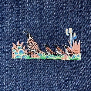 May include: Embroidered patch featuring a desert scene with a quail and three chicks. The patch has a light brown, white, and black quail with a cactus and other desert plants. The patch is on a blue denim background.