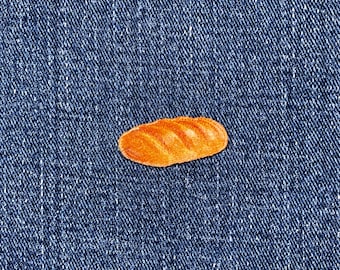 May include: A small, orange-brown bread loaf sticker on a blue denim background. The sticker has a detailed texture, with visible ridges and a slightly glossy finish. The denim has a classic, textured appearance.