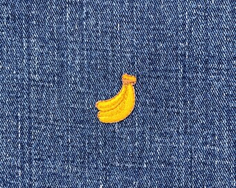 May include: A yellow banana patch with brown detailing is set against a blue denim backdrop. The embroidered patch shows a curved banana shape, with a textured look. The denim fabric has a visible weave.