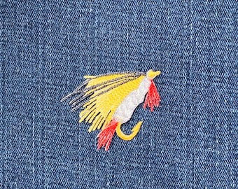 May include: Embroidered fishing fly patch in yellow, white, and red on a blue denim background. The fly has a curved hook and detailed stitching, suggesting a handcrafted item. The design is centered, showcasing the fly's intricate details.