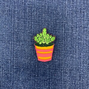 May include: An embroidered patch featuring a green cactus with multiple arms planted in a pink and yellow striped pot. The cactus has dark green soil at the base and is stitched with light green thread to show texture and detail on the plant.