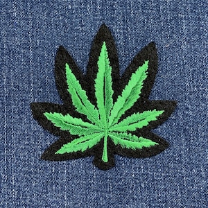 May include: Embroidered patch with a green cannabis leaf design, outlined in black. The patch is displayed on a blue denim background, highlighting the detailed stitching and bright colours. Suitable for jackets, bags, or other fabric items.