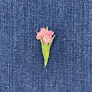 May include: An embroidered patch featuring a pink iris flower with a yellow center and green stems and leaves, stitched onto a dark blue denim fabric background. The floral design displays detailed thread work and vibrant colors.