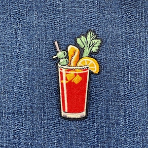 May include: An embroidered patch featuring a tall glass filled with a red liquid, garnished with two green olives on a pick, a bright orange peel, a slice of lemon, and a stalk of green celery, all set against a dark blue denim fabric background.