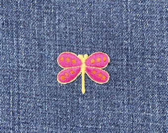 May include: A pink and orange embroidered dragonfly patch. The dragonfly has pink wings with orange spots and a yellow body. The patch is on a blue denim background.